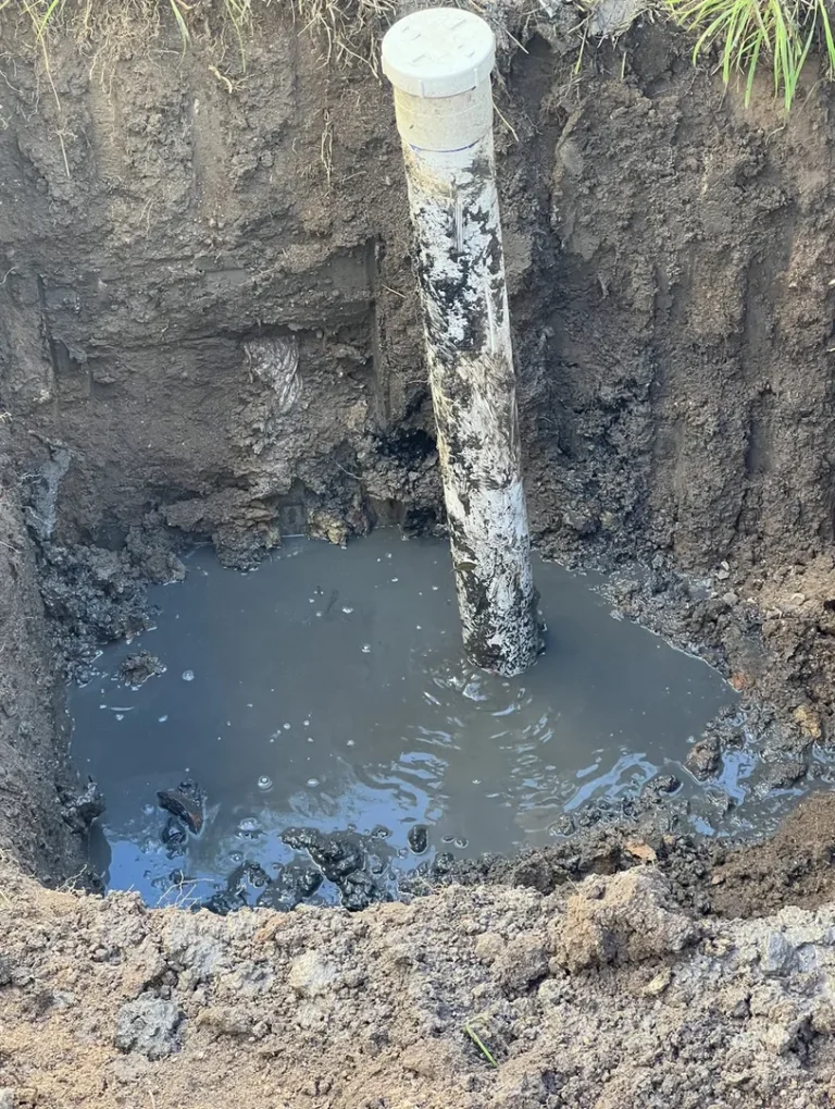 Excavated muddy hole with exposed PVC pipe.